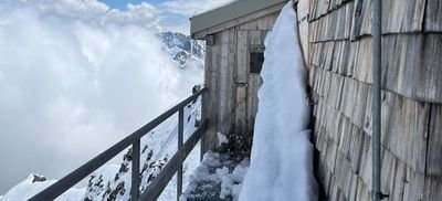 Wooden cabin in snowy mountains with snowdrifts, clouds, and mountain peaks in the background.