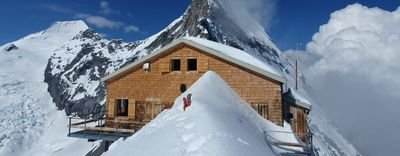 Wooden cabin in snowy mountains, person in red jacket standing on snow, blue sky in the background.
