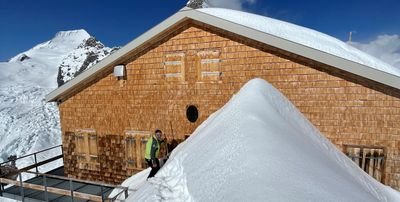 Person vor Berghütte im Schnee, umgeben von verschneiten Bergen, bei sonnigem Wetter.