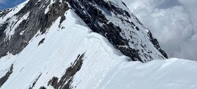 Verschneiter Berggipfel in den Alpen mit steilen Felswänden und Wolken im Hintergrund.
