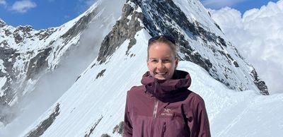 Person in lila Jacke steht vor schneebedecktem Berg in den Alpen, blauer Himmel im Hintergrund.