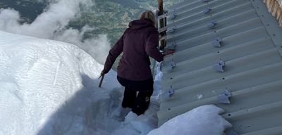 Person auf schneebedecktem Berg neben Hütte, hält sich an Geländer fest.
