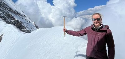 Person on snow-covered mountain with sunglasses and ice axe, surrounded by clouds.