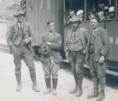 Four men in traditional clothing stand next to a train, surrounded by mountains.