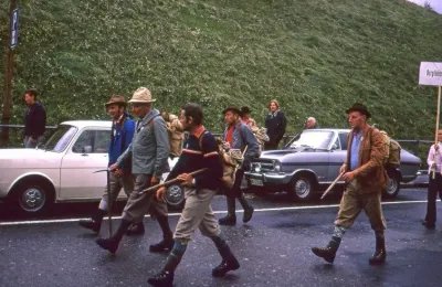 Group of people in traditional clothing with hiking sticks on a road in front of cars.