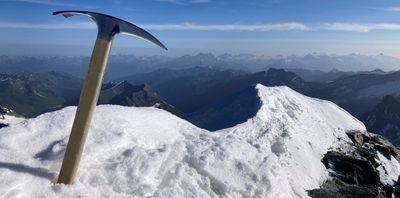Ein Eispickel steckt im Schnee auf einem Berggipfel mit Blick auf eine Berglandschaft.