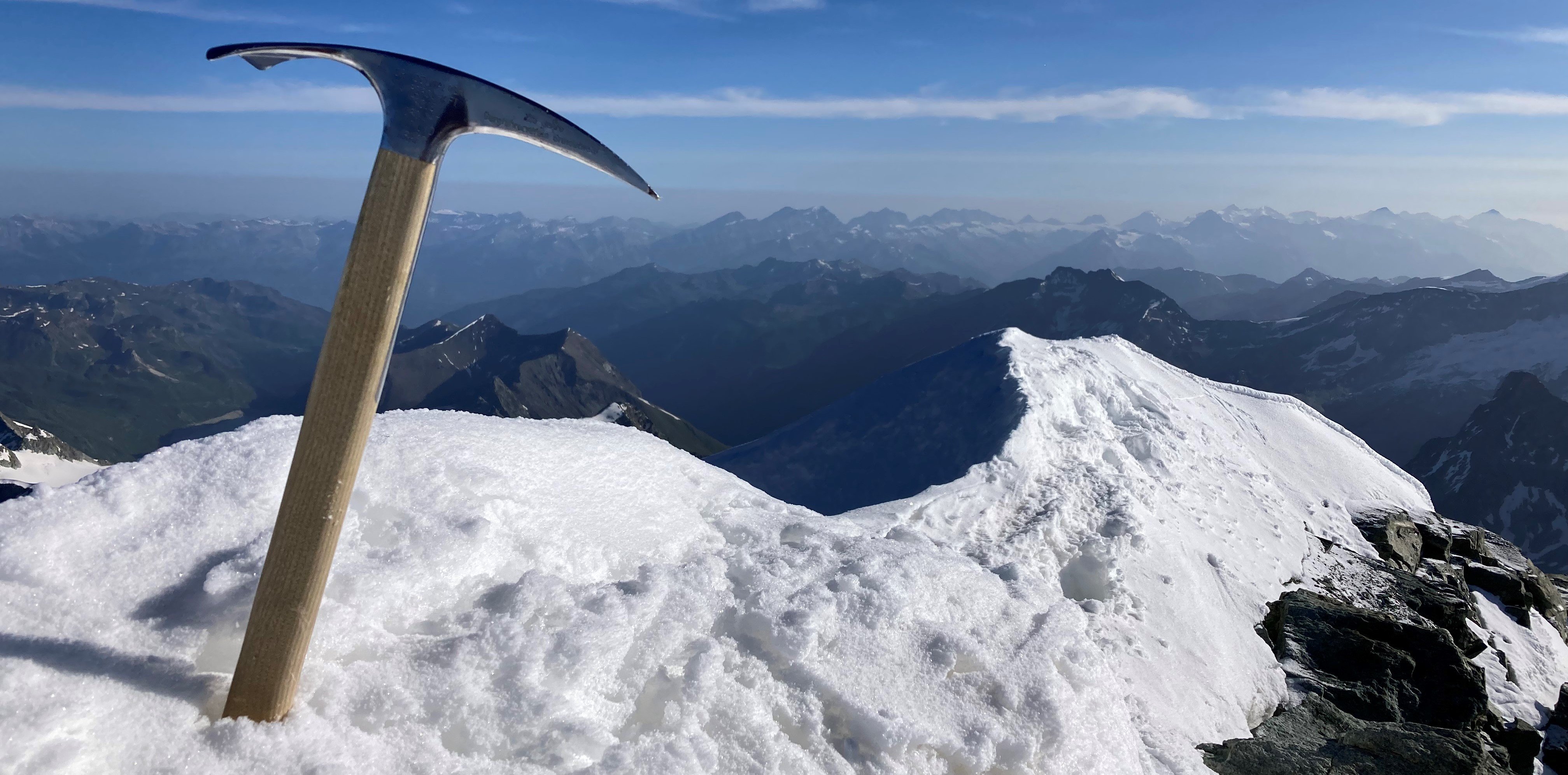 An ice axe is stuck in the snow on a mountain peak overlooking a mountain landscape.