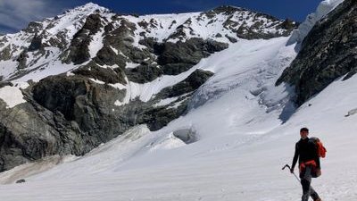 Person with ice pick and backpack on snow-covered mountain in front of rocky backdrop.