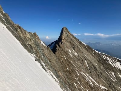 Berggipfel mit schneebedecktem Hang und felsiger Spitze, umgeben von blauen Himmel und fernen Bergen.