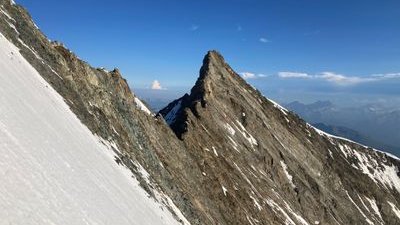 Mountain peak with a snow-covered slope and rocky summit, surrounded by blue sky and distant mountains.
