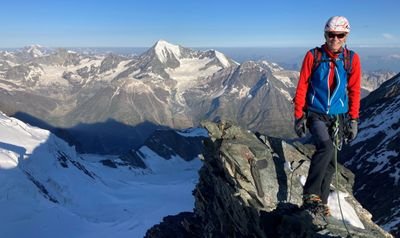 Person mit Helm und Kletterausrüstung steht auf Berggipfel, umgeben von schneebedeckten Alpen.
