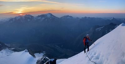 Bergsteiger mit Helm und Seil auf schneebedecktem Hang, im Hintergrund Berge bei Sonnenaufgang.