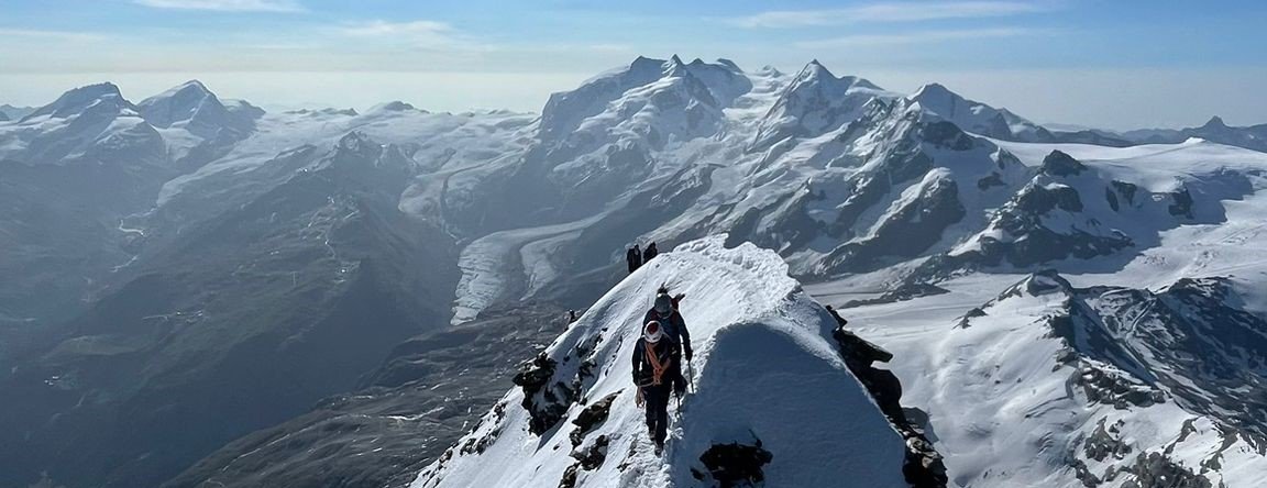 Mountaineers with helmets and ropes on a snow-covered ridge in the Alps, surrounded by mountains.