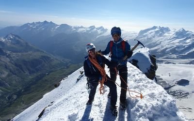 Two people with helmets and climbing gear are standing on a snow-covered mountain peak.