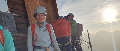People with helmets and climbing harnesses in front of a wooden hut in a mountain landscape at sunrise.