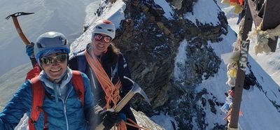 Two people with helmets and ice axes mountaineering on a snow-covered peak in the Alps.