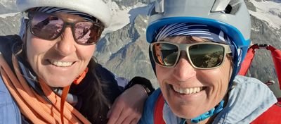 Two people with helmets and sunglasses in front of snow-covered mountains, one of them carrying a rope.