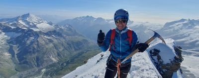 Person with helmet and ice pick on snow-covered mountain peak, with the Alps panorama in the background.