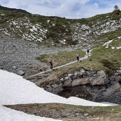 Im foreground there is a snow field and behind it a snow-free hiking trail with some people.