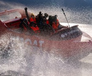 People in life jackets on a red speedboat, splashing through water, with mountains in the background.