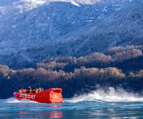 A red jet boat speeds across a lake with passengers in life jackets, set against snowy mountains.