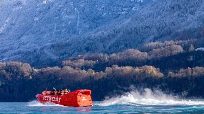 A red jet boat speeds across a lake with passengers in life jackets, set against snowy mountains.