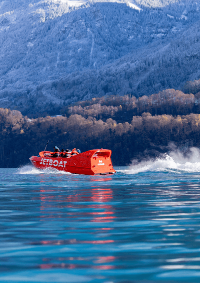 Rotes Jetboot mit Personen auf einem See, umgeben von bewaldeten Bergen und schneebedeckten Gipfeln.