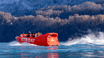 Rotes Jetboot mit Personen auf einem See, umgeben von bewaldeten Bergen und schneebedeckten Gipfeln.