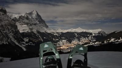 Schneeschuhe im Vordergrund, Berglandschaft und beleuchtetes Tal im Hintergrund bei Nacht.