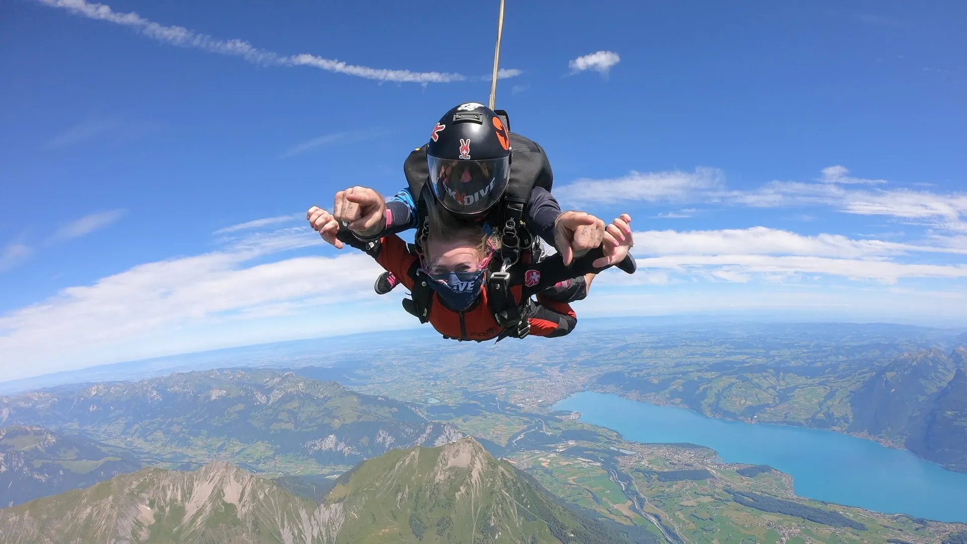 Two people tandem skydiving with helmets and harnesses over a mountain landscape with a lake.