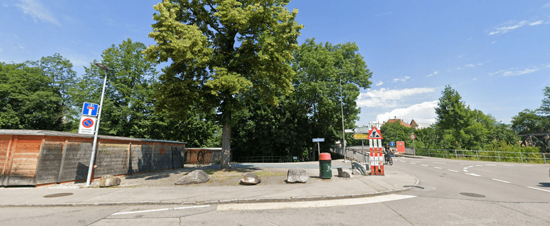 Leere Straßenszene mit Verkehrsschildern, Bäumen, Holzzaun und einem Mülleimer unter einem blauen Himmel.