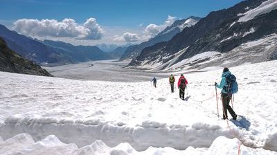 Hikers with backpacks and ropes traverse a snowy mountain landscape under a clear sky.