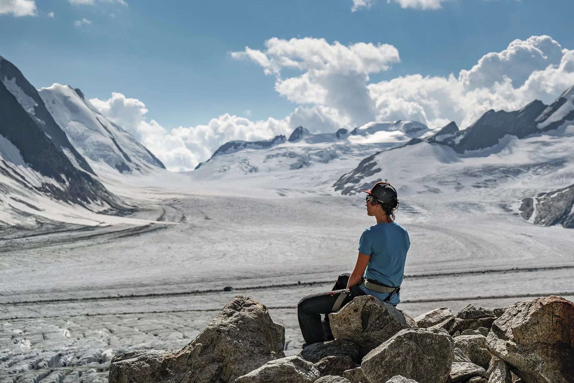 Person in helmet and harness sits on rocks, overlooking snowy mountains and glacier under cloudy sky.