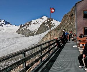 People in outdoor gear on a mountain terrace with a glacier view and Swiss flag.