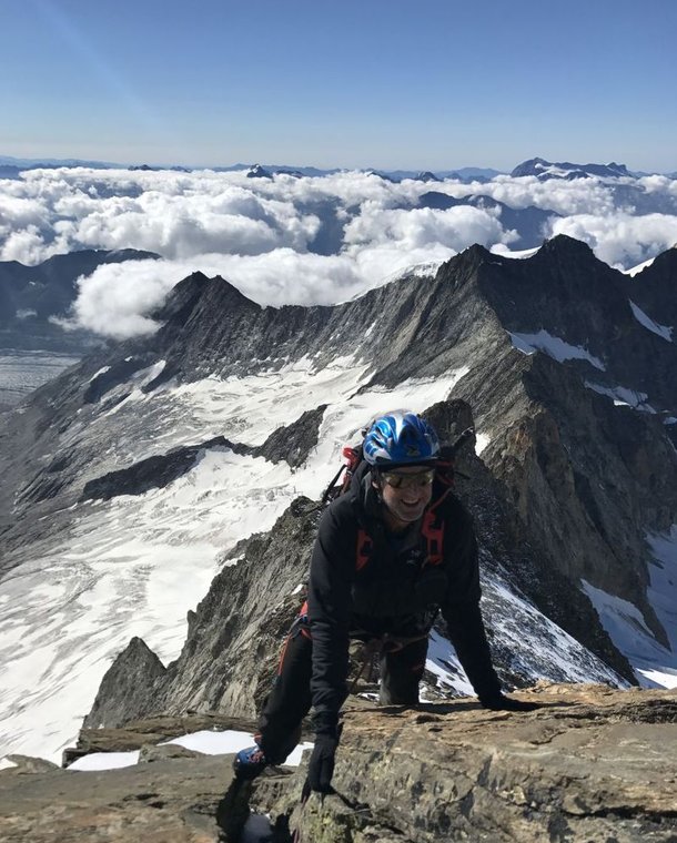 Climber with helmet and harness ascends rocky mountain ridge, snowy peaks and clouds in background.