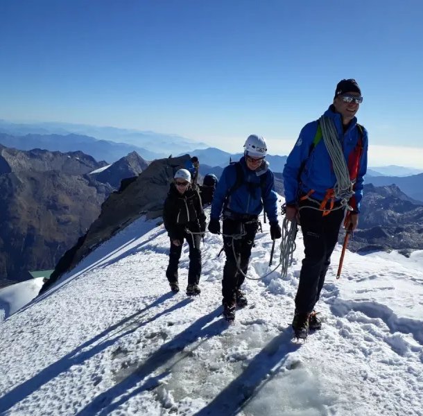 Three climbers with helmets and ropes ascend a snowy mountain ridge with a scenic view in the background.