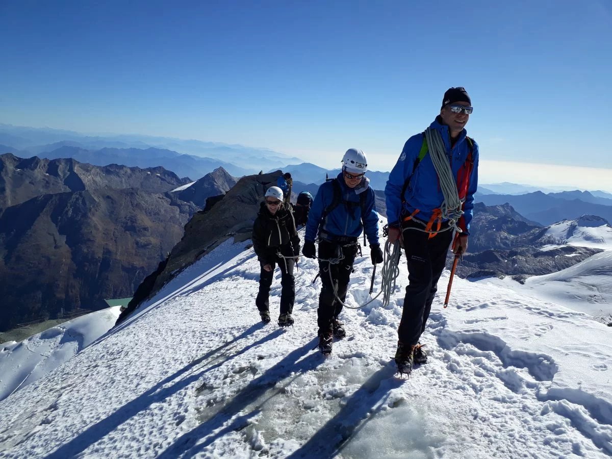 Three climbers with helmets and ropes ascend a snowy mountain ridge with a scenic view in the background.