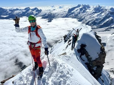 Bergsteiger mit Helm und Kletterausrüstung auf schneebedecktem Grat in alpiner Berglandschaft.