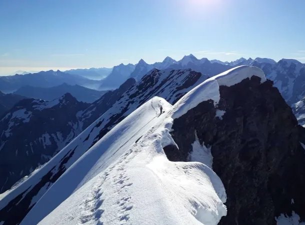 Climber with a helmet and harness traverses a snowy mountain ridge under clear skies.