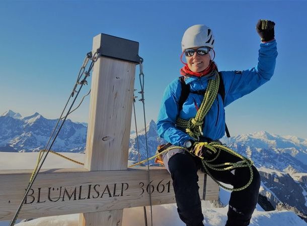 Climber in helmet and harness sits on summit with rope, snowy mountains in background.