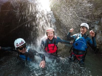 Drei Personen lachen unter einem Wasserfall beim Canyoning in einer Schlucht.