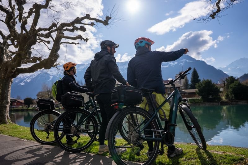 Three people with helmets and bicycles at the lakeshore, mountains in the background, sunny weather.