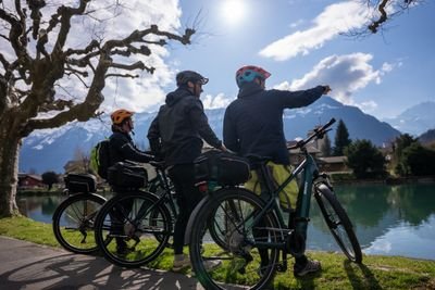 Three people with helmets and bicycles at the lakeshore, mountains in the background, sunny weather.