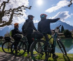 Three people with helmets and bicycles at the lakeshore, mountains in the background, sunny weather.