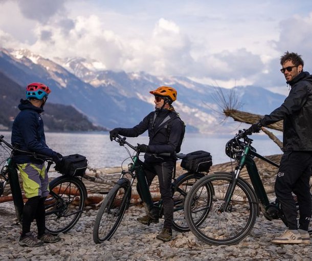 Three people with helmets on e-bikes at the lakeshore, surrounded by mountains and clouds.