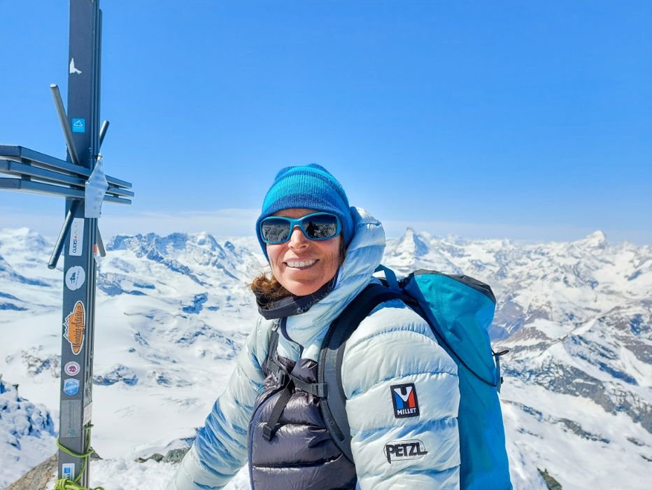 Person with a blue cap and sunglasses on a snow-covered mountain peak next to a summit cross.