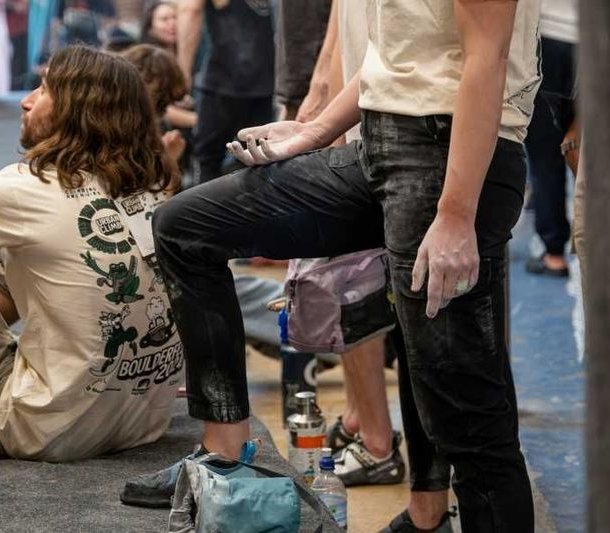 People in climbing gym with chalk on hands, wearing casual clothing, climbing equipment visible.