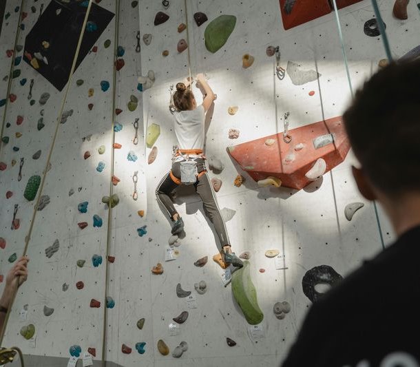 Person climbing on an indoor climbing wall with a harness, secured by another person.