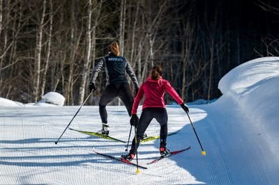 Two people cross-country skiing on a groomed snow trail, surrounded by trees.