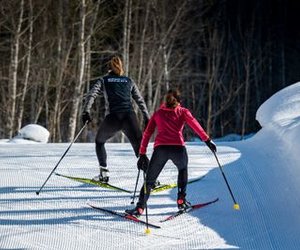 Zwei Personen beim Langlaufen auf einer präparierten Schneepiste, umgeben von Bäumen.